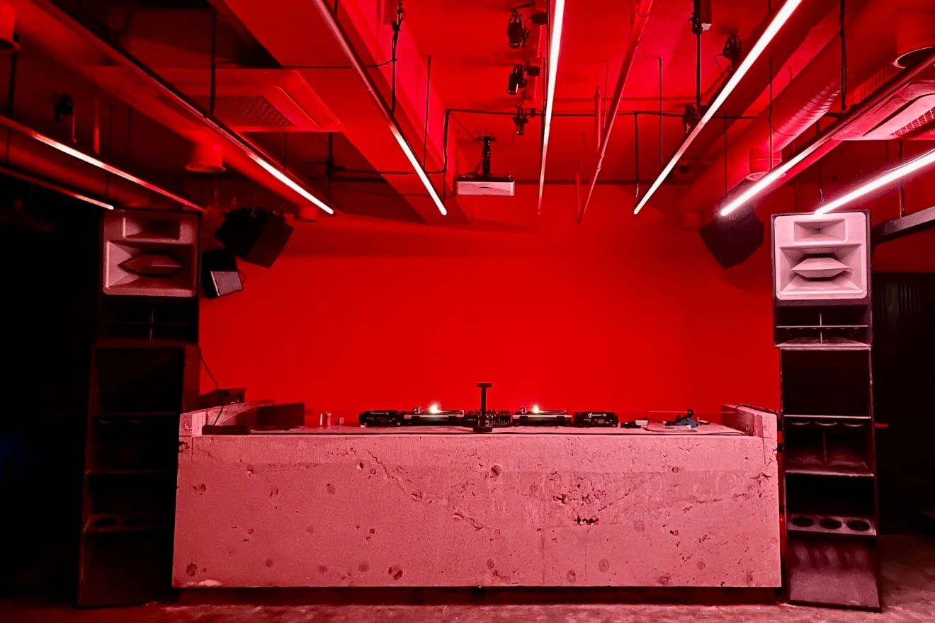 Archive view of TELLER’s empty DJ booth and speaker stacks beneath red ceiling lights.